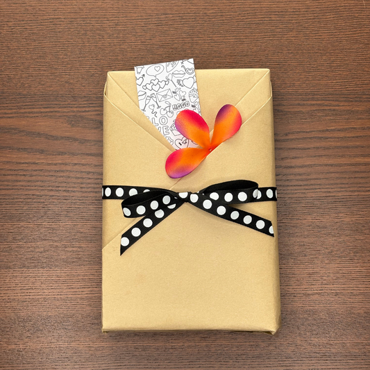This image showcases a charmingly wrapped book, ideal for bookworms and readers alike. The book is covered in eco-friendly kraft paper and tied with a black ribbon with white polka dots, giving it a cozy, bookish aesthetic. A black-and-white doodle bookmark peeks from the top, featuring reading-themed sketches like hearts, envelopes, and books. A faux flower in vibrant pink and orange adds a pop of color. Perfect for showcasing bookish merch, handmade gifts for readers, aesthetic book mail, or creative book